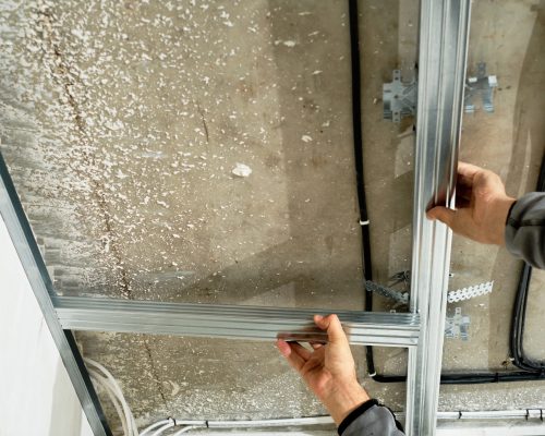 Worker adjusts a metal profile for mounting a plasterboard ceiling frame, close-up, selective focus on the hands of a specialist. Industrial renovation and renovation